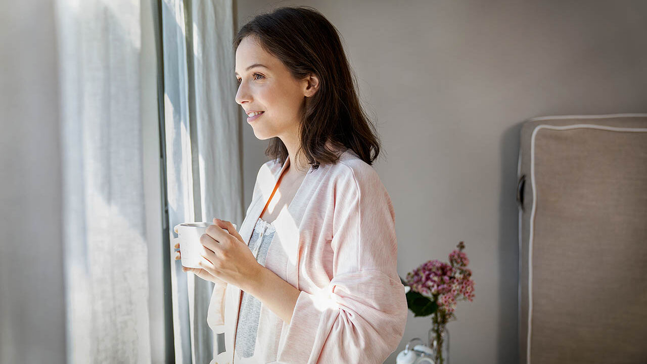 Eine Frau steht morgens in einem Bademantel und mit einer Tasse Tee am Fenster und sieht hinaus.