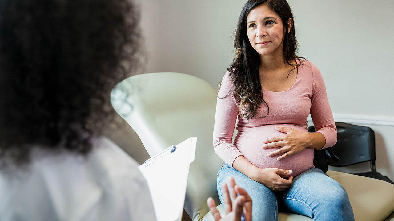 Eine schwangere Frau sitzt auf einer Behandlungsliege einer Arztpraxis und wird von einer Ärztin beraten.