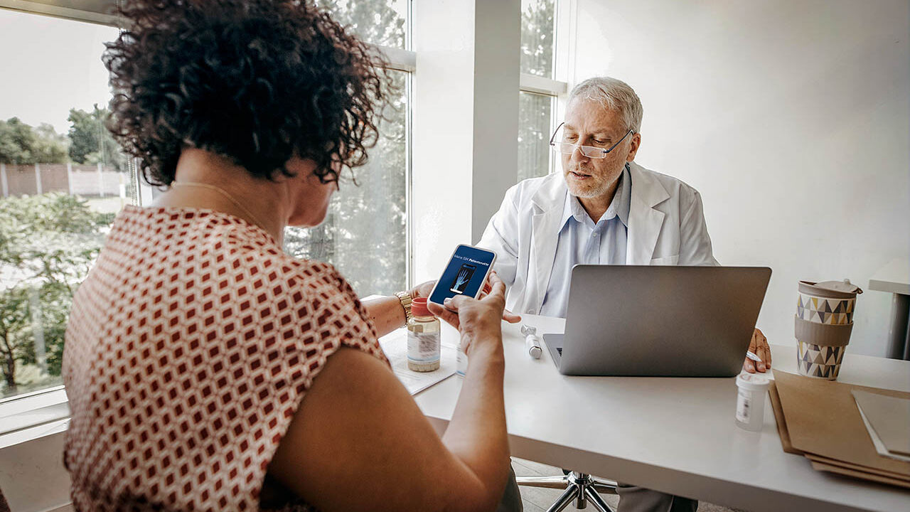 Eine Frau sitzt mit einem Arzt an einem Beratungstisch und teilt ein Dokument über ihre elektronische Patientenakte mit ihm. ihm.