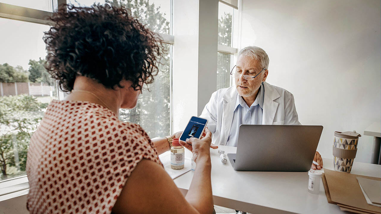 Eine Frau sitzt mit einem Arzt an einem Beratungstisch und teilt ein Dokument über ihre elektronische Patientenakte mit ihm. ihm.