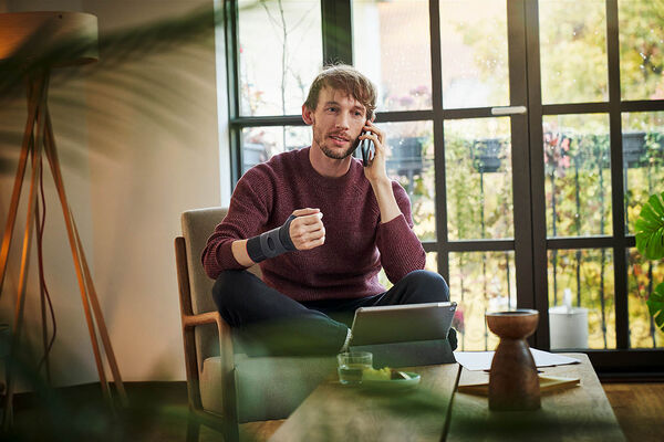 Ein Mann sitzt mit einer Schiene am Handgelenk zu Hause im Wohnzimmer und benutzt mit der anderen Hand sein Handy.