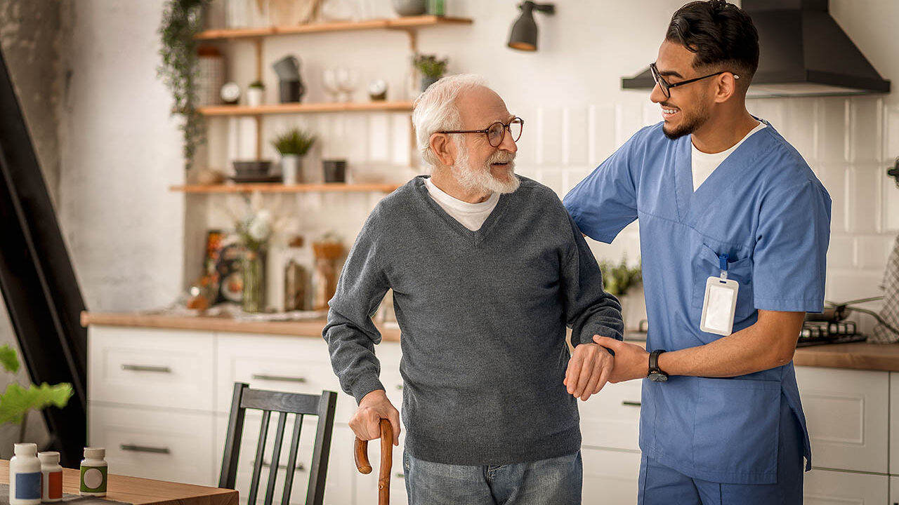 Ein  älterer Mann wird von einem Pfleger beim Laufen gestützt.