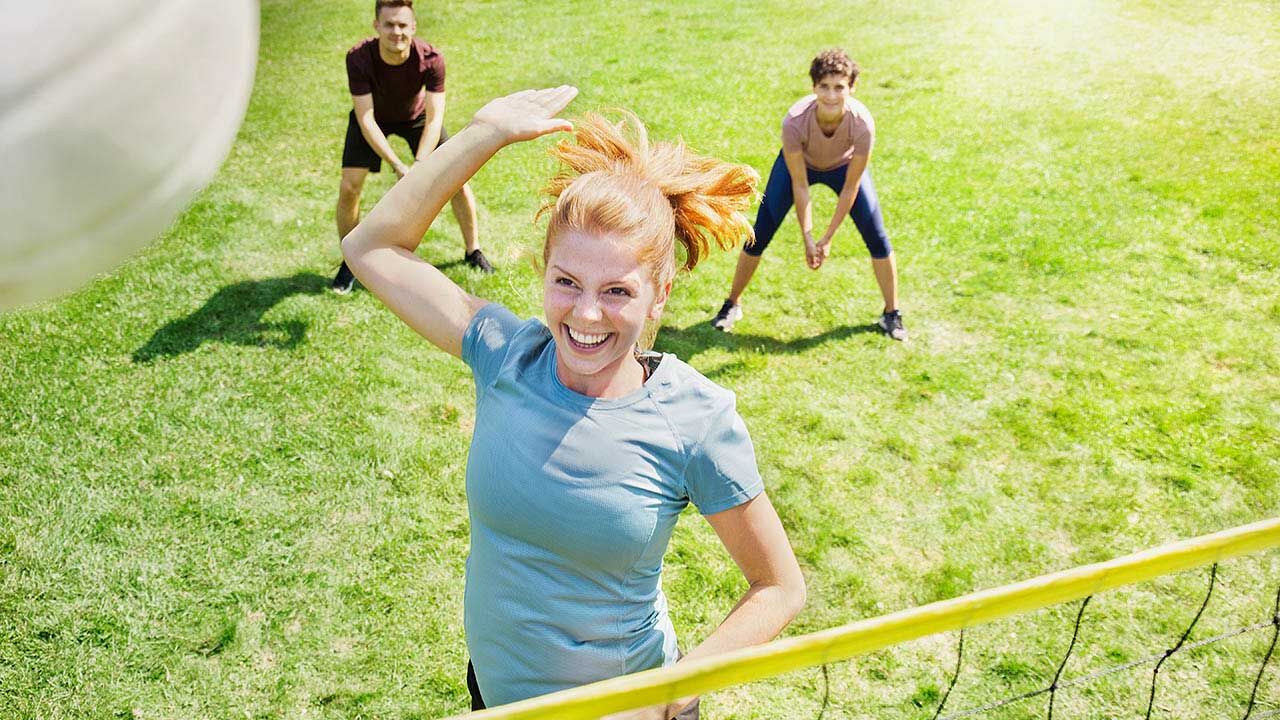 Eine Frau spielt mit Freunden im Park Volleyball und springt gerade hoch, um den Ball über das Netzt zu schlagen..