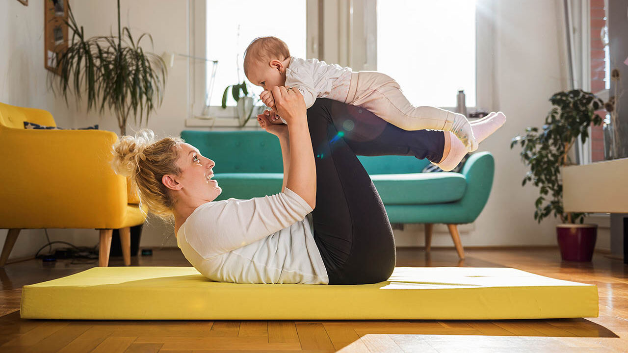 Eine Mutter macht Rückbildungsübungen mit ihrem Neugeborenen auf einer Sportmatte im Wohnzimmer.