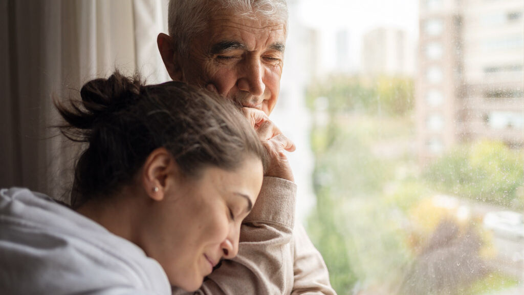 Ein Mann steht nachdenklich am Fenster. Eine junge Frau lehnt sich liebevoll an ihm an.