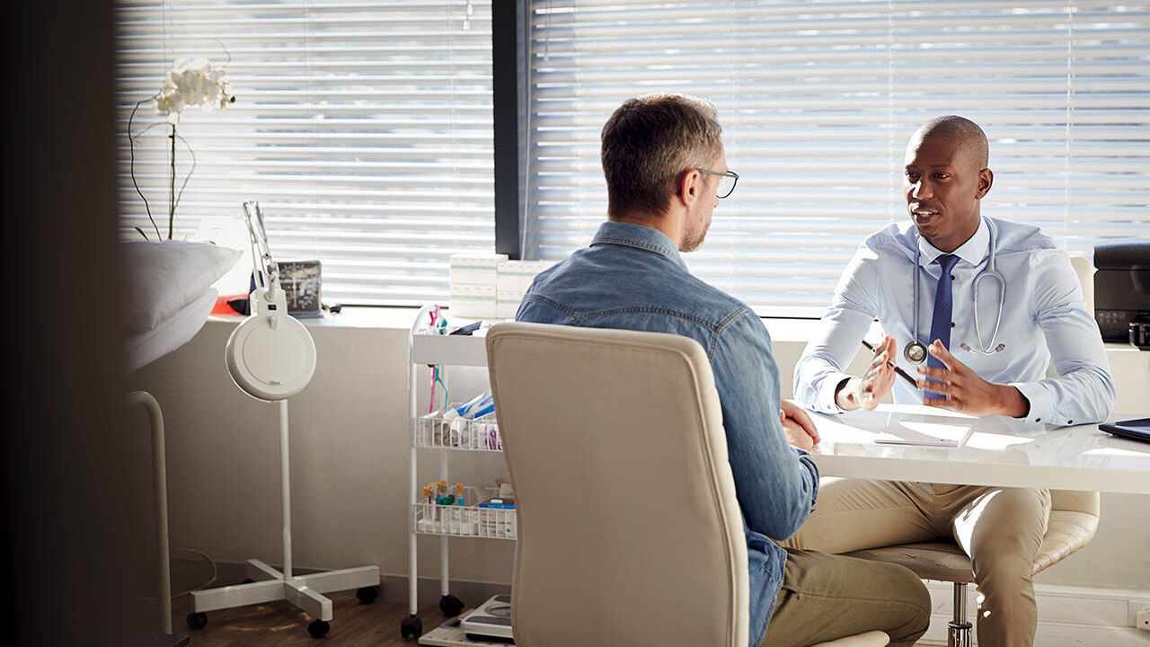 Ein Patient sitzt in einem Behandlungsraum seinem Arzt gegenüber und sie besprechen sich.