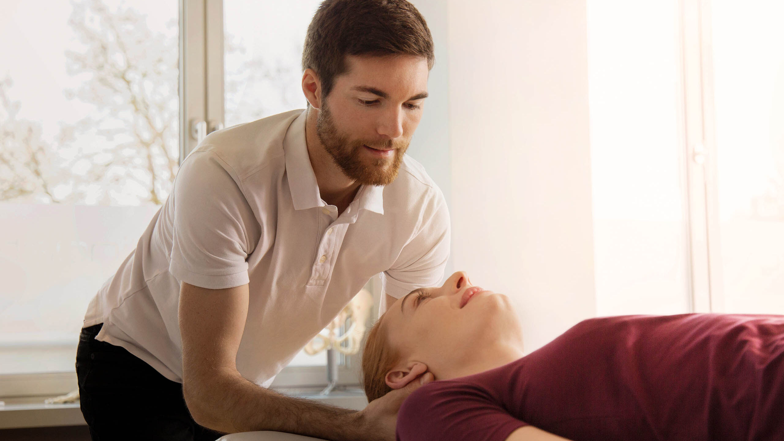 Ein Osteopath untersucht und behandelt den Nacken einer Patientin, die auf einer Liege im Behandlungsraum liegt.