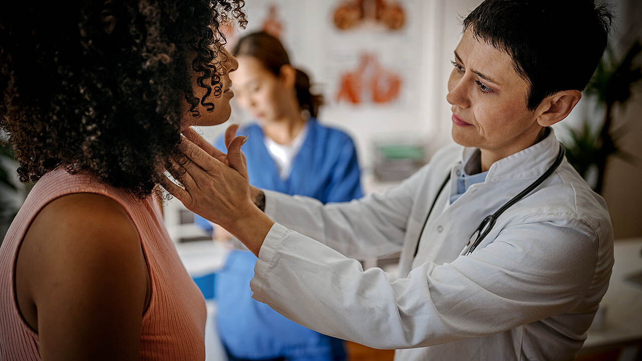 Eine Ärztin untersucht eine junge Frau an ihrem Hals mittels einer Tastuntersuchung.