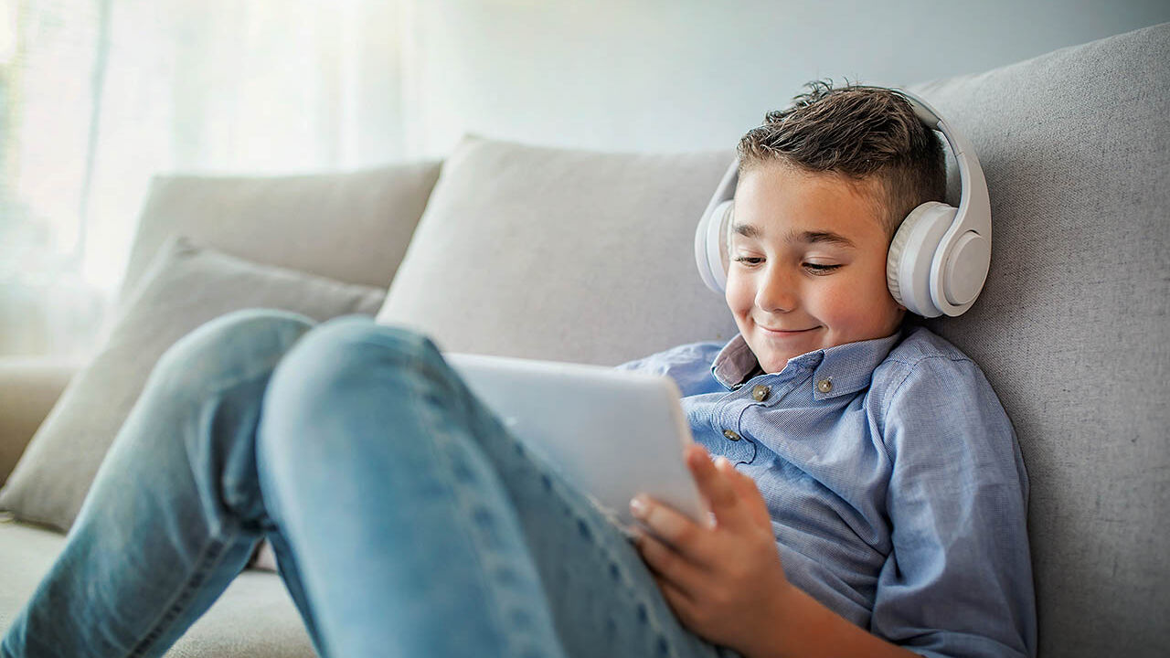 Ein Junge sitzt mit Kopfhörer und einem Tablet auf dem Sofa und lächelt.