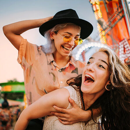 Zwei junge Frauen sind auf einem Festplatz und lachen miteinander.