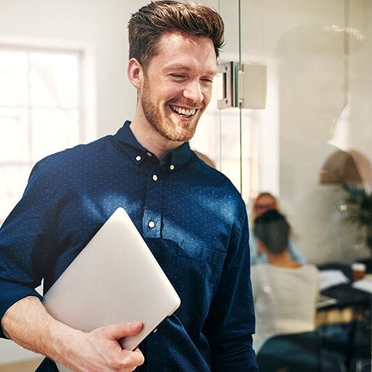 Ein junger Mann im Büro mit Laptop unter dem Arm.