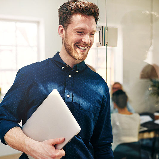Ein junger Mann im Büro mit Laptop unter dem Arm.