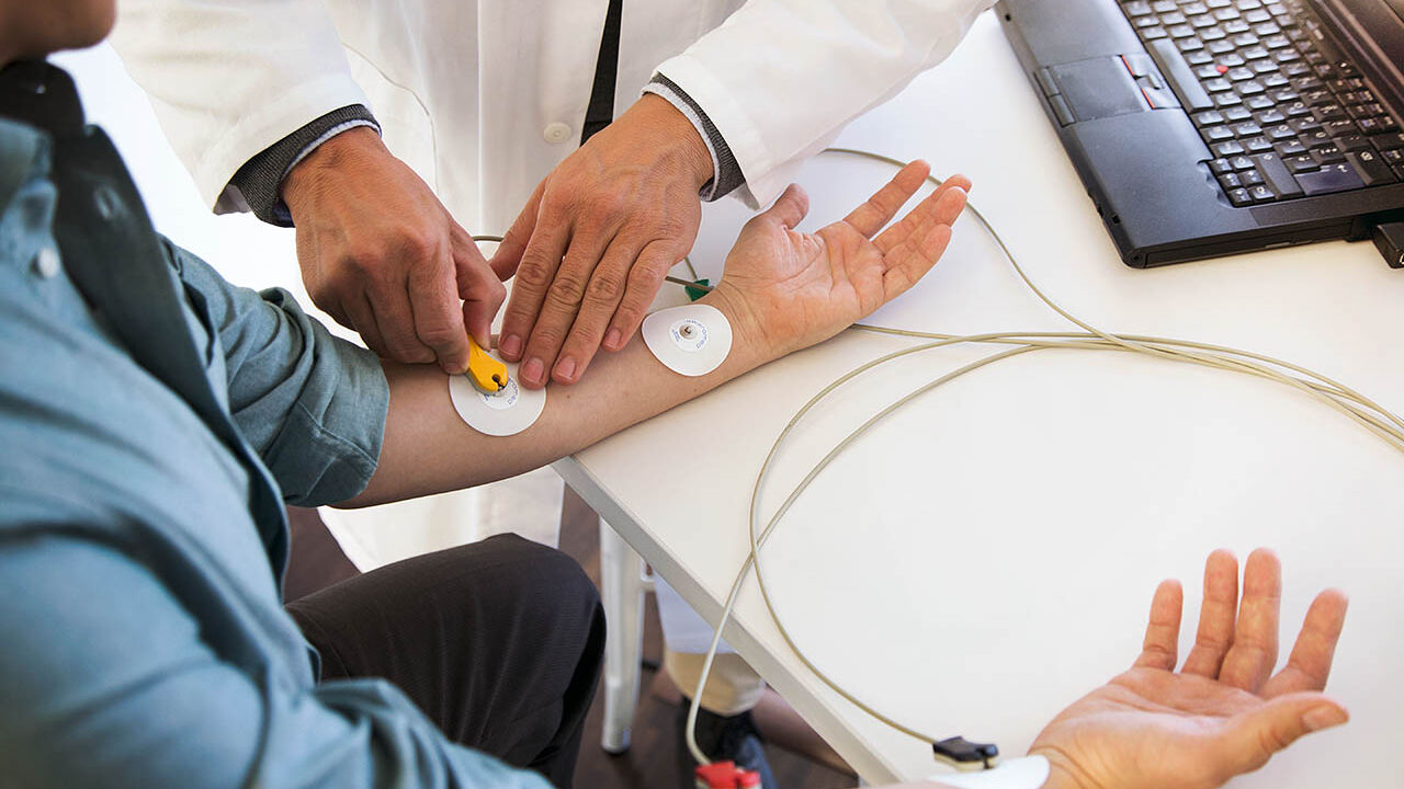 Ein Mann sitzt im Behandlungszimmer und ein Arzt platziert Elektroden eines EKG-Geräts auf seinen Unterarmen.