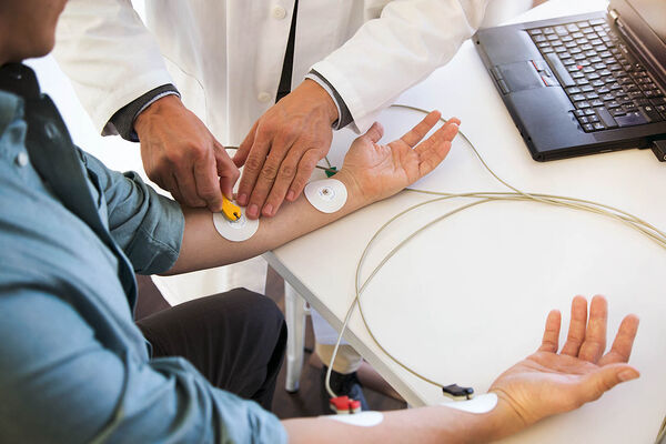 Ein Mann sitzt im Behandlungszimmer und ein Arzt platziert Elektroden eines EKG-Geräts auf seinen Unterarmen.