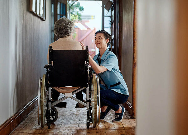 Eine Pflegefachkraft sitzt in der Hocke neben einer älteren Frau im Rollstuhl und berührt sie an der Schulter.
