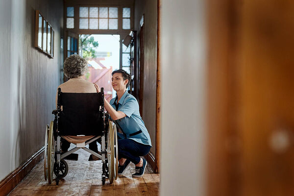 Eine Pflegefachkraft sitzt in der Hocke neben einer älteren Frau im Rollstuhl und berührt sie an der Schulter.