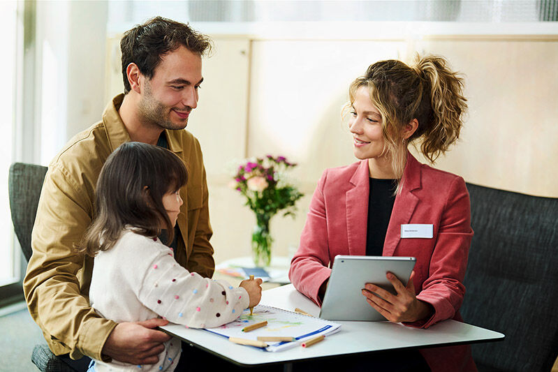 Eine Mitarbeiterin der SBK sitzt mit einem Versicherten und seiner kleinen Tochter an einem Tisch und berät sie.