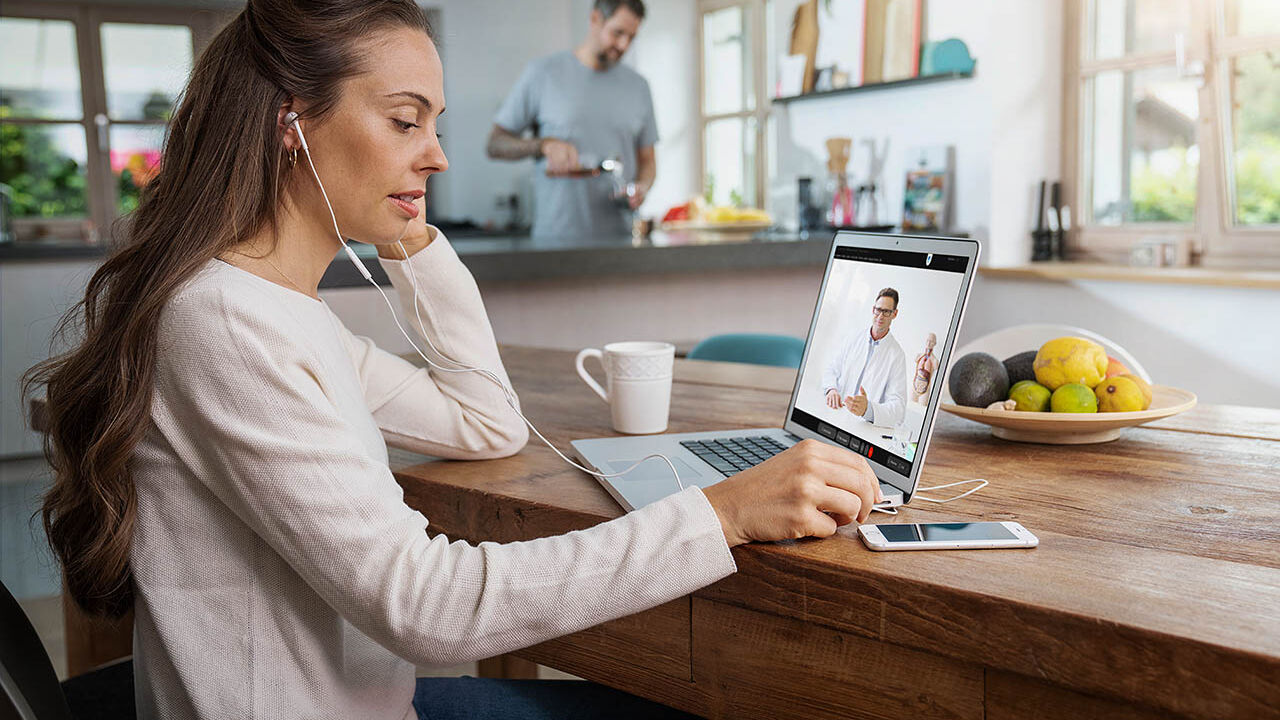 Eine Frau sitzt an ihrem Esstisch und hat über ihren Laptop gerade eine Videoberatung mit einem Arzt.