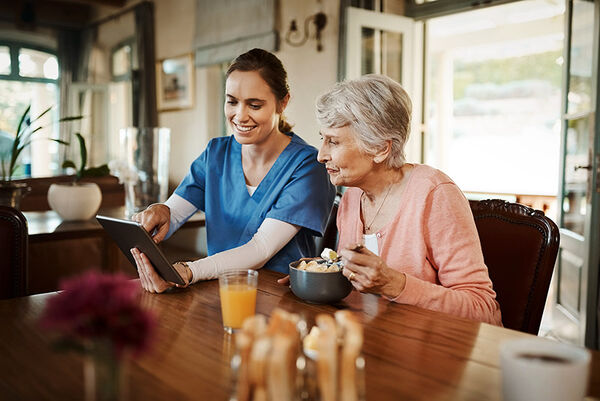 Eine Pflegefachkraft sitzt mit einer älteren Frau am Esstisch und zeigt ihr etwas auf dem Tablet.