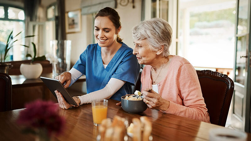 Eine Pflegefachkraft sitzt mit einer älteren Frau am Esstisch und zeigt ihr etwas auf dem Tablet.