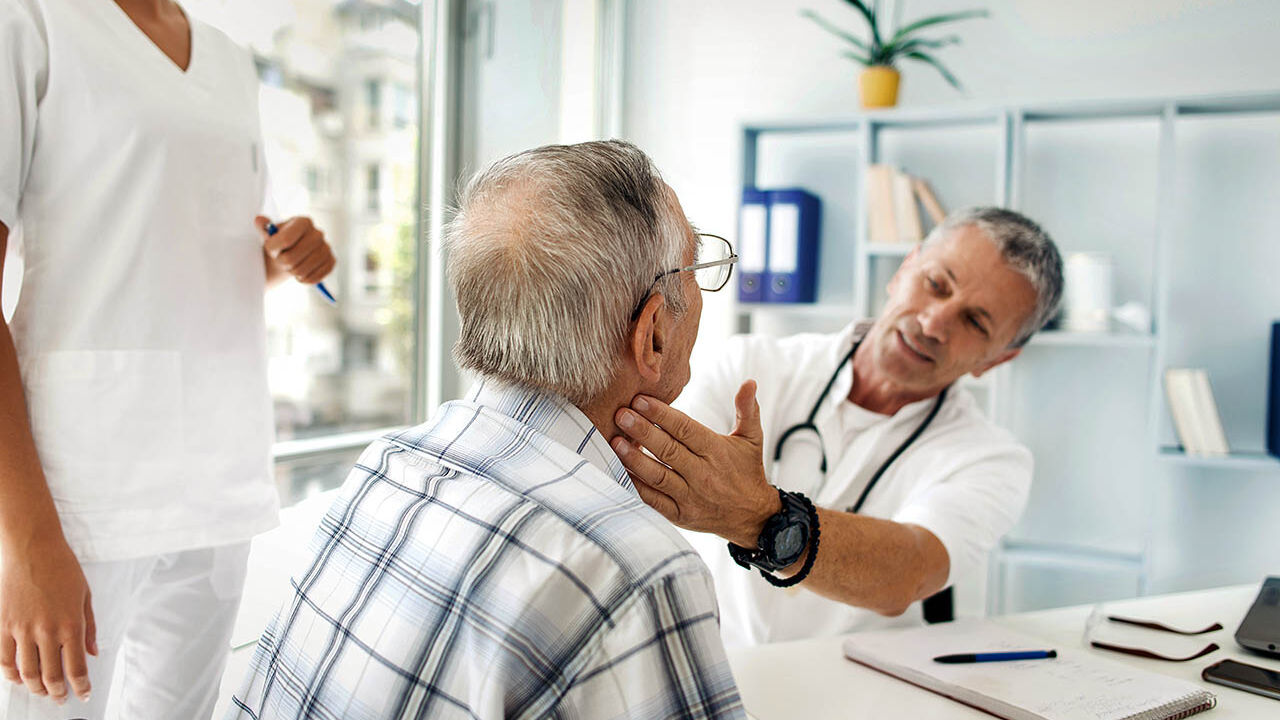 Ein Arzt untersucht in seinem Behandlungszimmer einen älteren Mann durch eine Tastuntersuchung am Hals.