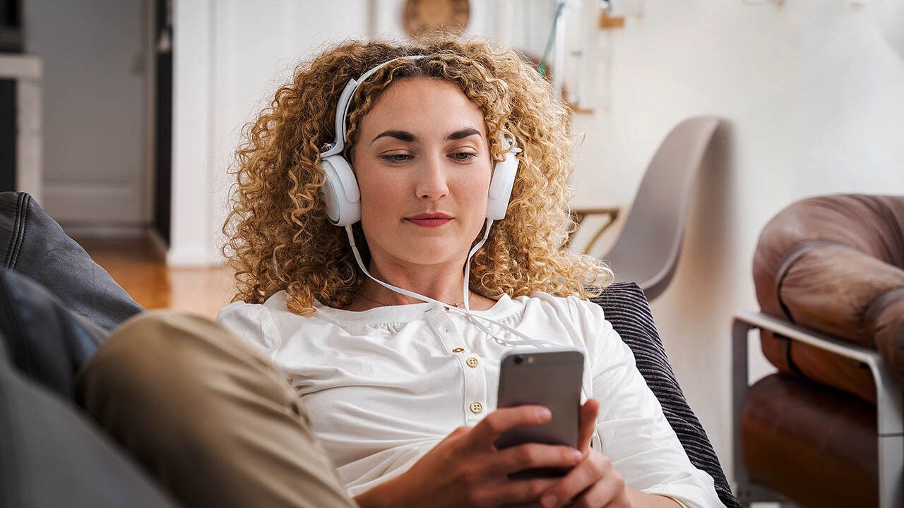 Eine Frau sitzt mit Kopfhörern auf dem Sofa und schaut auf ihr Handy.