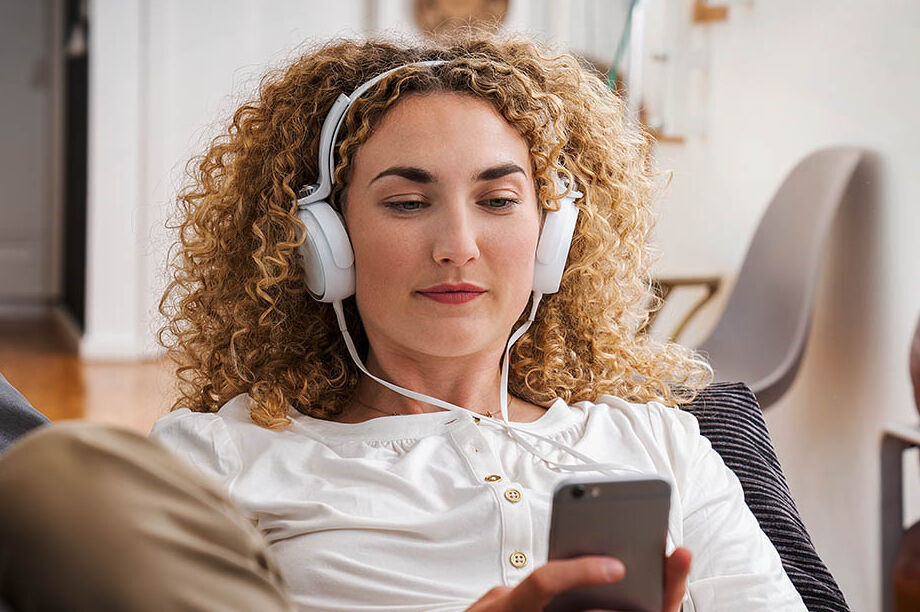 Eine Frau sitzt mit Kopfhörern auf dem Sofa und schaut auf ihr Handy.