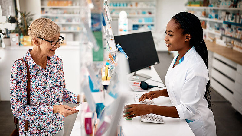 Eine Frau löst ihr Arzneimittelrezept in einer Apotheke ein und wird von einer Apothekerin über den Tresen beraten.