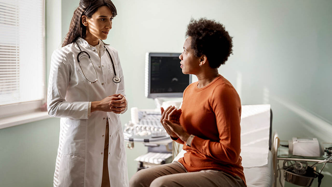 Eine Patientin sitzt auf einer Liege im Behandlungsraum und erklärt ihre Symptome der Ärztin.