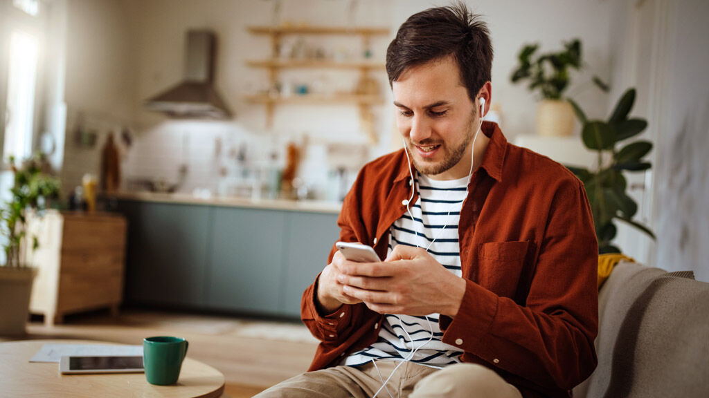 Ein Mann sitzt auf dem Sofa und hört etwas über Kopfhörer und sein Mobiltelefon an.