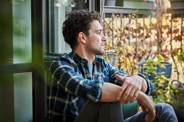 Ein Mann sitzt auf dem Boden an seine Terassentür gelehnt und sieht nachdenklich auf den Boden vor sich.
