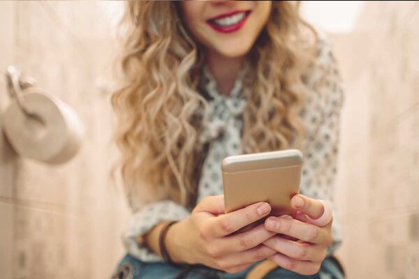 Junge Frau, die mit Handy in der Hand auf der Toilette sitzt