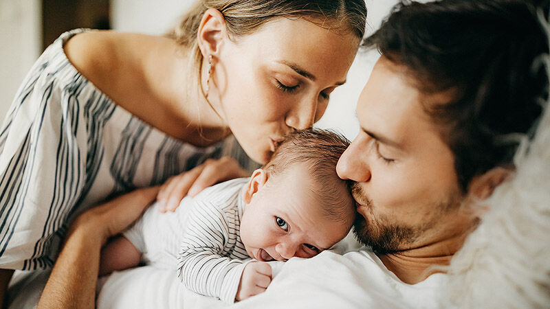 Ein Mann hat ein Neugeborenes auf sich liegen und er und eine Frau geben ihm ein Kuss auf den Kopf.