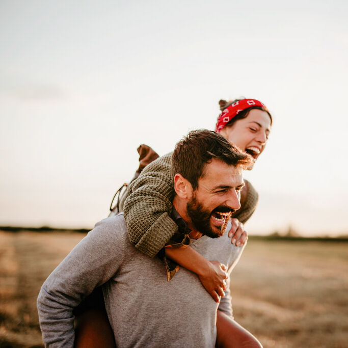 Ein Mann trägt eine Frau im Huckepack auf dem Rücken. Die beiden sind in der Natur und lachen herzlich.