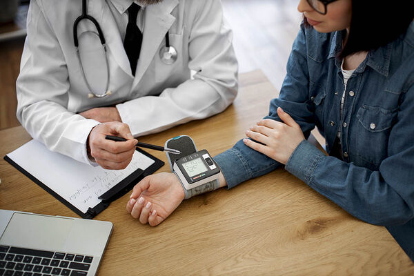 Ein Arzt und eine Patientin sitzen gemeinsam an einem Tisch und messen mit einem Gerät an ihrem Handgelenk ihren Blutdruck.