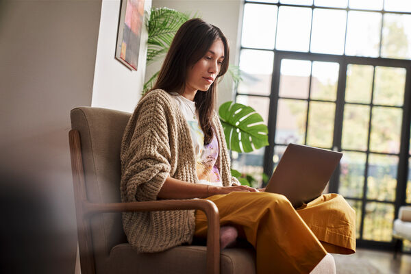 Eine Frau sitzt in ihrer Wohnung auf einem Sessel und blickt auf den Laptop auf ihrem Schoß.