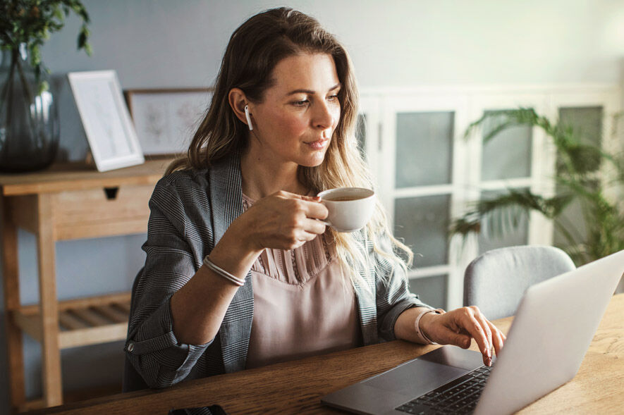 Eine Frau sitzt mit Kopfhörer und einer Tasse Tee vor ihrem Laptop im Esszimmer.