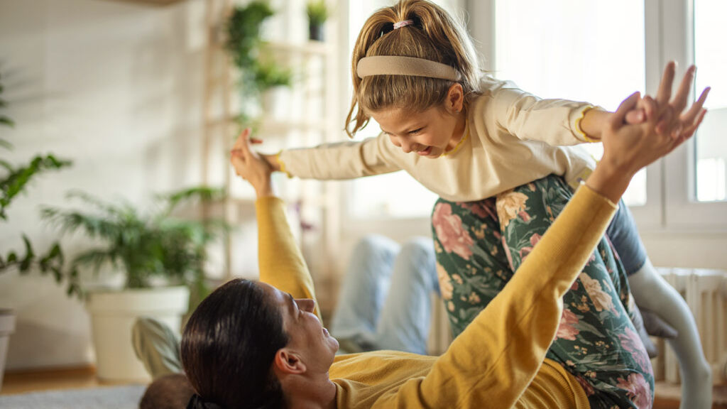 Eine Frau liegt auf dem Rücken auf dem Boden und balanciert ihre Tochter spielend auf ihren Knien.