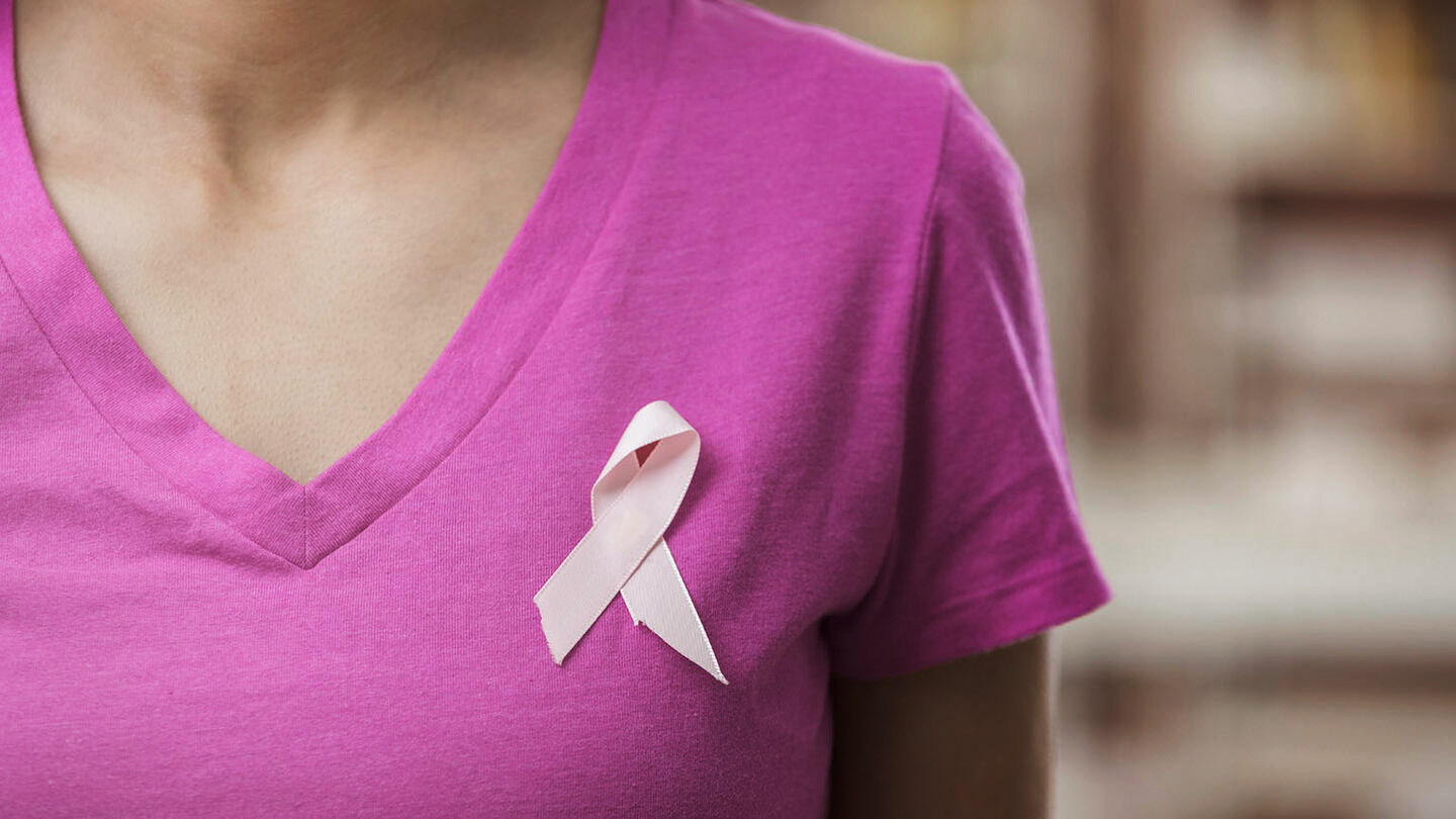 Eine Frau trägt ein rosa farbiges Shirt, woran eine Brustkrebs-Schleife befestigt ist.