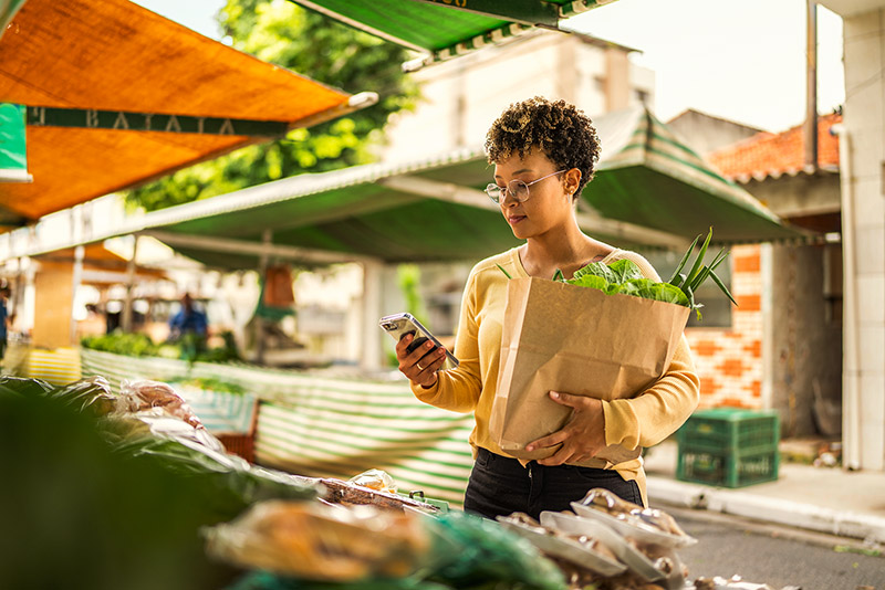 Eine Frau steht an einem Marktstand, hält Gemüse im Arm und schaut auf ihr Smartphone.