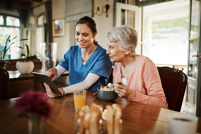 Eine Pflegefachkraft sitzt mit einer älteren Frau am Esstisch und zeigt ihr etwas auf dem Tablet.