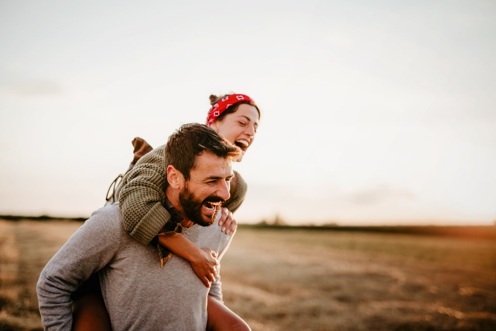 Ein Mann trägt eine Frau im Huckepack auf dem Rücken. Die beiden sind in der Natur und lachen herzlich.
