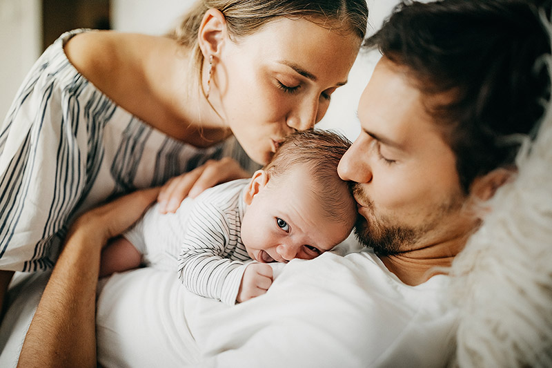Ein Mann hat ein Neugeborenes auf sich liegen und er und eine Frau geben ihm ein Kuss auf den Kopf.