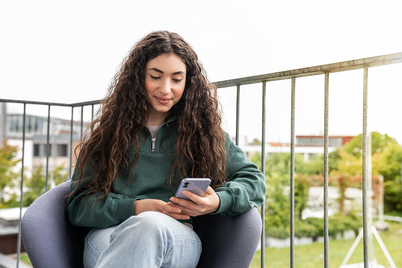 Ein Mädchen sitzt auf dem Balkon und schaut in ihr Handy.