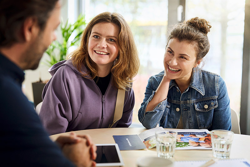 Zwei junge Frauen sitzen mit einem SBK-Kundenberater an einem Tisch in einer Beratungszone und lachen gemeinsam.